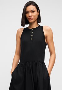 Μαύρο αμάνικο φόρεμα με εφαρμοστή κορυφή, στρογγυλή λαιμόκοψη και λεπτομέρεια κουμπιών, φαρδύ φούστα και πλαϊνές τσέπες. Λείος υφής.