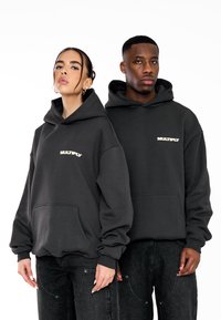 Sudaderas gris oscuro con un bolsillo frontal, que presentan la palabra "MULTIPLICAR" en un beige claro. Corte suelto con capuchas grandes y puños acanalados.