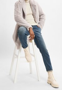 Abrigo de piel sintética gris claro sobre un suéter crema, combinado con jeans de mezclilla azul y botas beige con cordones, sentado en un taburete blanco.