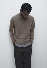 Un pull en tricot marron avec un col zippé quart, porté sur une chemise bleu clair, associé à un pantalon foncé. Texture lisse et coupe décontractée.