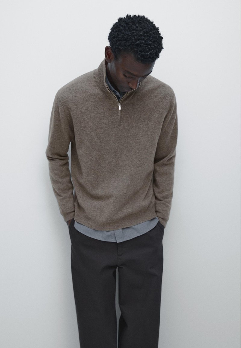 Un pull en tricot marron avec un col zippé quart, porté sur une chemise bleu clair, associé à un pantalon foncé. Texture lisse et coupe décontractée.
