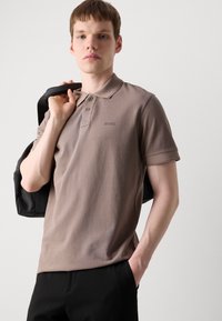 Brun polo-skjorte med korte ermer, tre-knaps placket og preget logo på brystet. Modellen holder en svart veske over den ene skulderen.