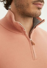 Pull-over rose clair avec un col montant, doté d'une fermeture éclair demi-longueur et d'un intérieur gris. Texture lisse avec un détail de tirette en cuir.