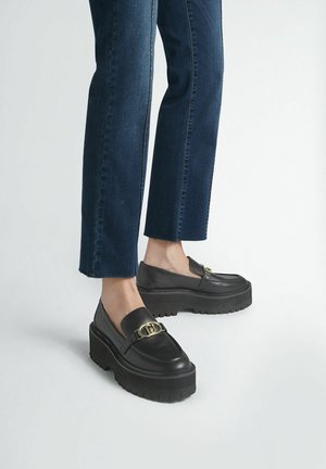 Mocassini in pelle nera con suola piatta spessa e dettaglio in fibbia dorata, abbinati a pantaloni cropped in denim scuro con orli sfrangiati.