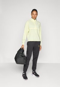 Ljusgul halvdragkedja jacka, svarta joggers med logotyp, kombinerat med svarta sneakers och en svart duffelväska. Släta texturer, atletisk passform.
