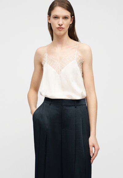 Γυναίκα που φοράει λευκό καμιζολάκι με δαντέλα, φορεμένο μέσα σε ψηλόμεσο, πιετέ μαύρο παντελόνι, στέκεται μπροστά σε ένα απλό φωτεινό φόντο.