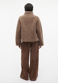 Bruine nepbontjas met een hoge kraag en brede mouwen, gecombineerd met hooggeknoopte bruine suède broek. Beide kledingstukken hebben een strakke afwerking.
