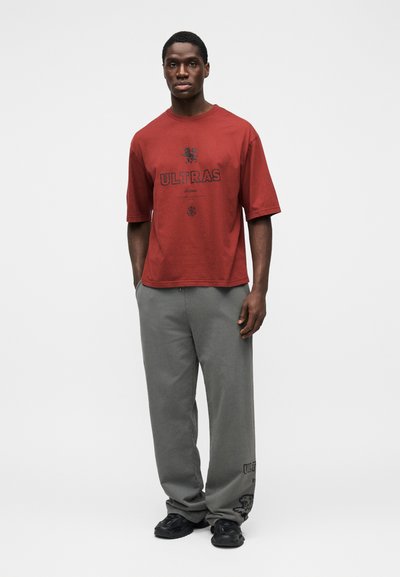 T-shirt rouge en coton avec un motif noir "ULTRAS", assorti à un pantalon ample gris avec un accent lion sur l'ourlet et des baskets noires.