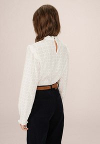 Blusa bianca con un motivo texture, colletto arricciato e dettaglio a chiave sul retro; abbinata a pantaloni di corduroy scuri e una cintura marrone.