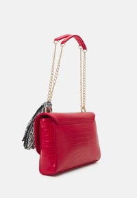 Borsa a tracolla in pelle sintetica rossa effetto coccodrillo con una tracolla in catena dorata e un elegante pom pom nero e bianco. Texture liscia, forma rettangolare.