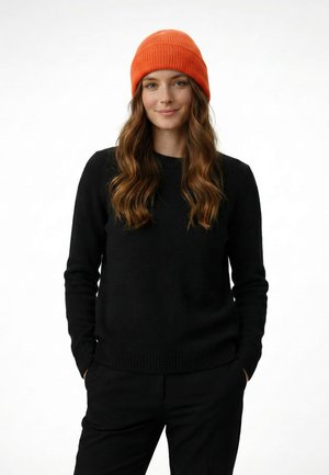 Young woman with long wavy hair wearing an orange knit beanie and black sweater, standing with hands in pockets, against white background.