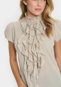 Camicia beige con design a balze e colletto alto. Maniche a sbuffo corte con chiusura a bottone. Tessuto leggero che offre un aspetto testurizzato.