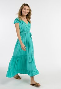 Rochie maxi turcoaz cu mâneci volan, talie ajustabilă și fustă în straturi. Purtați-o cu sandale embellizate, iar materialul pare neted și fluid.