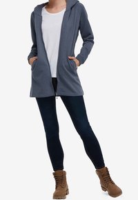 Felpa grigia con zip e tasche, indossata sopra una t-shirt bianca. Jeans skinny blu e stivali marroni con lacci completano l'outfit.