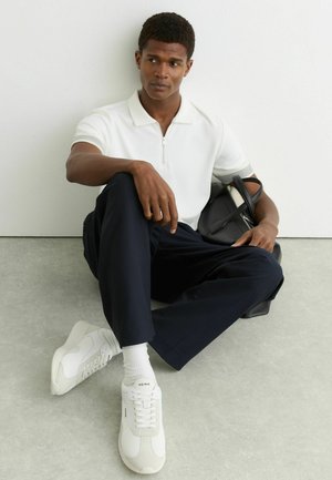 Sedící muž v bílém polo tričku, tmavě modrých kalhotách a bílých teniskách, držící černou tašku opřený o bílou zeď a šedou podlahu.