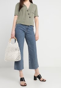 Grön kortärmad skjorta, blå korta jeans, svarta klacksandaler och en vit vävd väska med trähandtag.