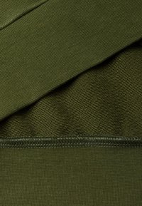 Tessuto verde oliva caratterizzato da sezioni texture e lisce, con cuciture visibili lungo i bordi. Composizione in materiale morbido e elastico.