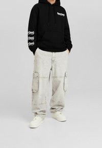 Persona che indossa un felpa nera con testo bianco sul petto e sulla manica, pantaloni cargo grigi chiari e sneaker bianche, con le mani in tasca, in piedi.