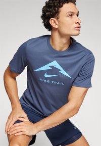 Tumed sinine Nike Trail T-särk, millel on helebeež mägine logo ja tekst. Lühikesed varrukad, ümmargune kaelus ja sile kangasteksts.
