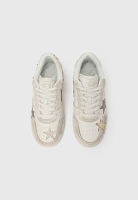 Baltas sporta kurpes ar mežģīņu aizdari, dekorētas ar pelēku un zeltu zvaigžņu uzšuvēm vienkāršā baltā fonā.