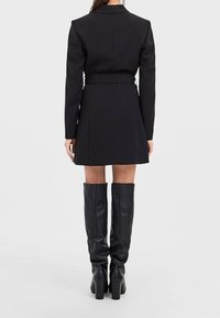 Kobieta ubrana w czarną sukienkę blazerową z paskiem, sięgającą do kolan czarne skórzane buty, stojąca tyłem na jednolitym tle.
