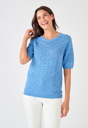 Donna sorridente con i capelli castani lunghi che indossa un maglione a maniche corte azzurro chiaro a motivo e pantaloni bianchi su uno sfondo semplice.