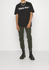 Zwarte graphic t-shirt met de tekst "vergeten gezichten", gecombineerd met olijfgroene joggingbroek en zwarte veterschoenen. Glad materiaal, losse pasvorm.
