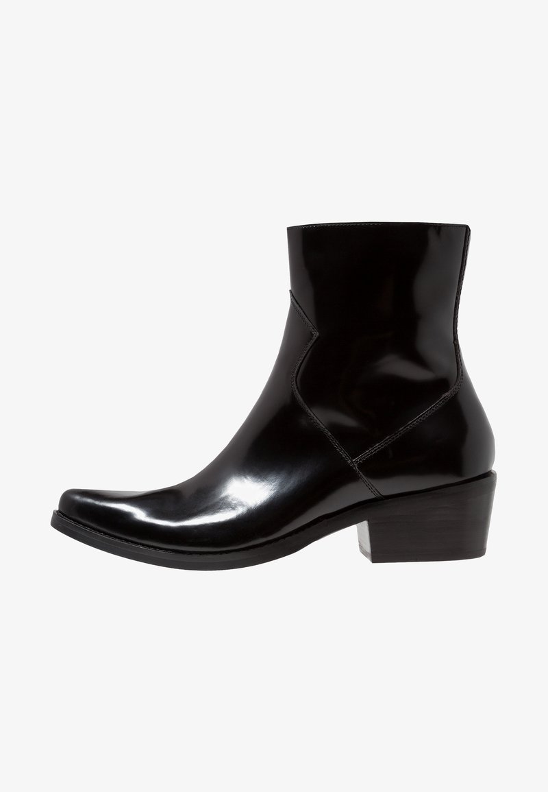 Bottine en cuir noir brillant avec bout pointu et petit talon bloc, détails de couture latérale, sur fond blanc.