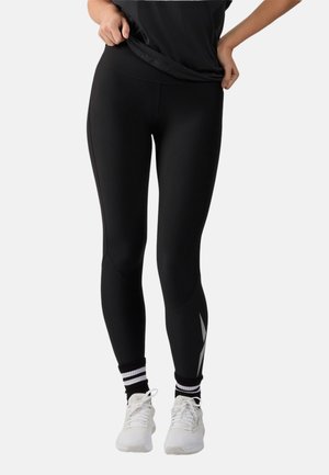 Vrouw draagt zwarte aansluitende legging met grijs patroon op de kuit, witte sneakers en zwarte sokken met witte strepen, tilt haar zwarte topje iets op.