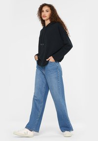 Schwarzer Kapuzenpullover mit Kordelzug, kombiniert mit hellblauen weiten Jeans. Das Model trägt weiße Sneakers mit hellbraunen Akzenten.