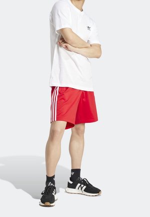 Persona in piedi con le braccia incrociate, indossa una t-shirt bianca Adidas, pantaloncini rossi Adidas con strisce bianche, calzini Adidas neri e scarpe da ginnastica nere.
