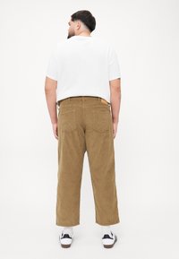 Pantalon en velours côtelé marron clair avec une coupe droite, présentant deux poches arrière et une étiquette beige sur la taille, associé à un t-shirt blanc.