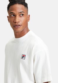 Biała bawełniana koszulka z okrągłym dekoltem, krótkimi rękawami i małym logo FILA w czerwonym i granatowym na lewej piersi.