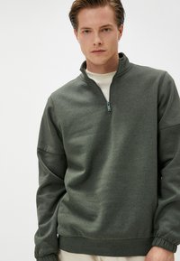 Sudadera verde oliva con cremallera hecha de tejido suave, con un alto cuello, mangas con puños y un ajuste relajado. Textura suave, diseño minimalista.