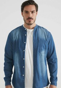 Homme aux cheveux courts et foncés portant une chemise en denim bleu déboutonnée sur un t-shirt blanc, se tenant devant un fond clair.