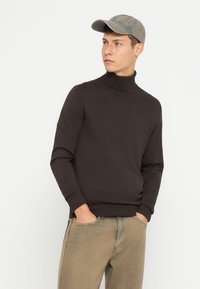 Bruine coltrui met lange mouwen, gemaakt van een gladde stof. Gecombineerd met lichtgroen-bruinachtige broek en een grijze pet.
