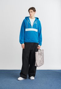 Jauns vīrietis, ģērbies spilgti zila Nike kapucē, platajās melnās biksēs, baltās kedās, turēt pelēku Nike somu, stāv uz zilā paklāja pret baltu sienu.