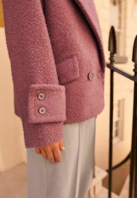 Blazer violet texturé avec boutons proéminents et une poche plaquée, associé à un pantalon gris clair. Il présente un matériau doux et duveteux.