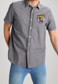 Korte mouwen grijze denim shirt met witte knopen, voorzien van een geborduurd tijgerhoofd op de linkerborst. Een gebogen zoomontwerp.