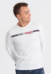 Weißes Langarm-T-Shirt mit roten und marineblauen Streifenakzenten und dem Text "OMBRE // MENSWEAR" auf der Vorderseite. Hergestellt aus weichem Baumwollstoff.