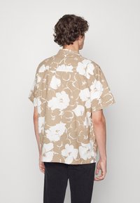 Camicia a maniche corte color beige con motivo floreale bianco. Realizzata in tessuto leggero, presenta un colletto classico e dettagli sul retro.