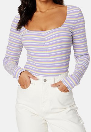 Mujer que lleva un top de manga larga a rayas en lavanda, amarillo y blanco con escote cuadrado y pantalones blancos de talle alto con las manos en los bolsillos.