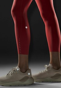 Czerwone legginsy sportowe o smukłym fasonie, z małymi otworami wzdłuż dolnej części, w zestawie z beżowymi, przewiewnymi sneakersami.