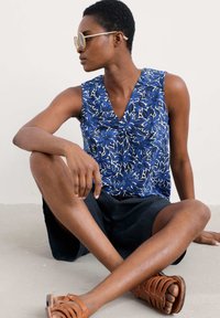 Blå blommig mönstrad ärmlös topp med V-ringning, i kombination med mörka shorts och bruna sandaler med remmar. Slätt tyg och avslappnad passform.