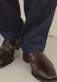 Brown leather boots with a polished finish, pointed toe, and low heel, paired with navy blue trousers featuring a textured fabric.