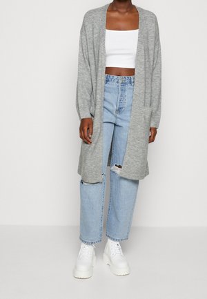 Personne portant un long cardigan gris, un crop top blanc, un jean droit bleu clair déchiré et des bottes blanches à plateforme sur un fond uni.