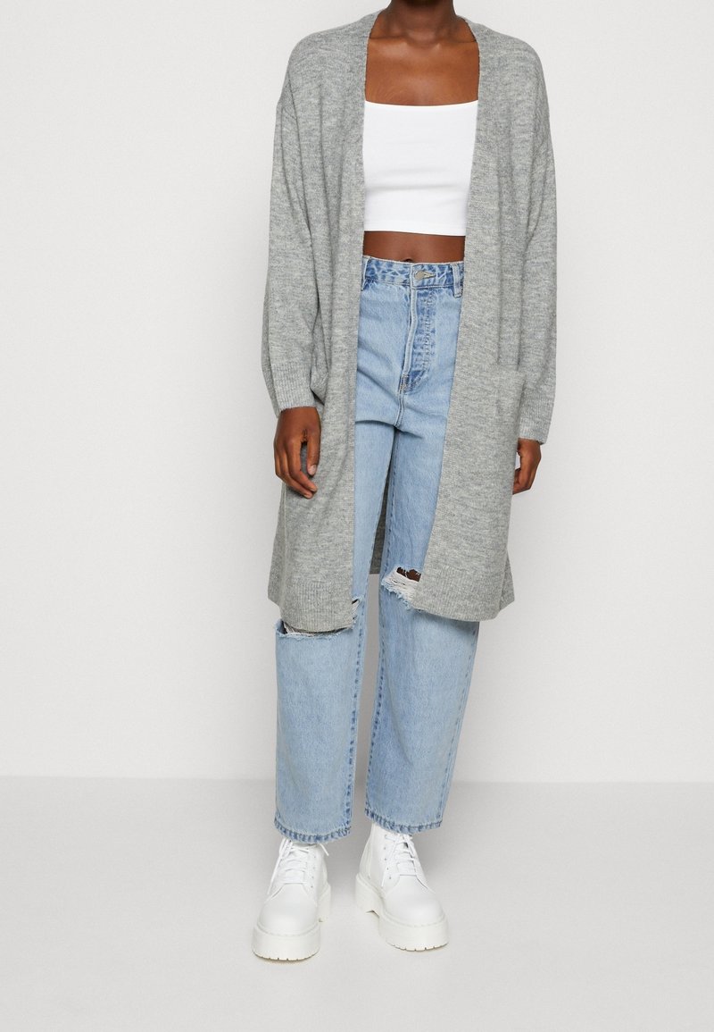 Personne portant un long cardigan gris, un crop top blanc, un jean droit bleu clair déchiré et des bottes blanches à plateforme sur un fond uni.