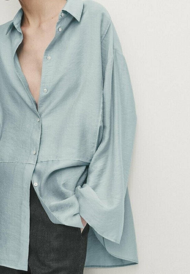 Personne portant une chemise bleu clair oversize à boutons avec manches larges et pantalon foncé, une main dans la poche, sur fond blanc uni.