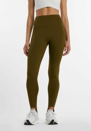 Donna che indossa leggings verde oliva a vita alta e sneakers bianche in piedi davanti a uno sfondo semplice.