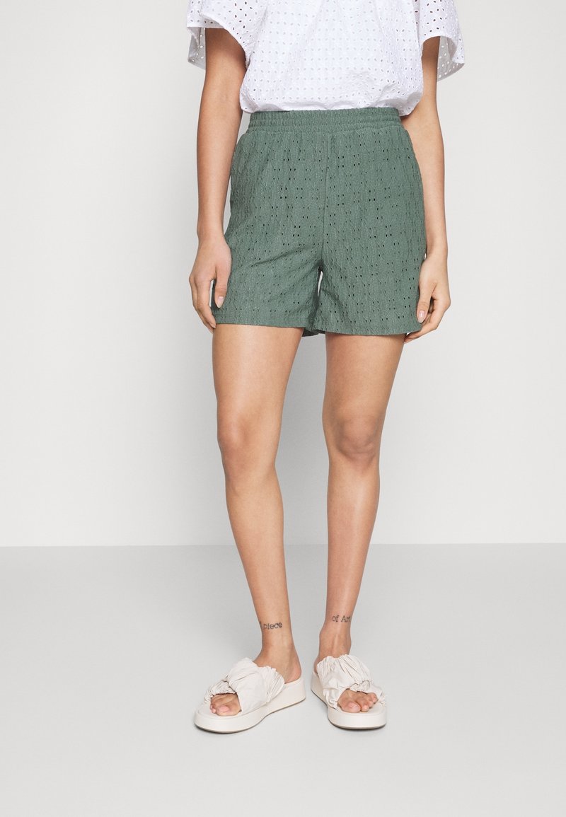Personne portant un short vert texturé, un haut blanc à œillets et des sandales blanches avec des brides volantées, debout devant un fond uni.